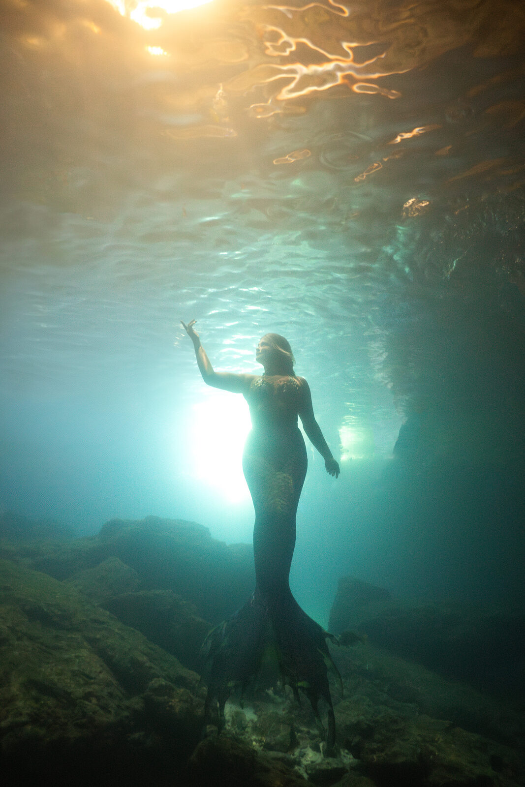 Mermaid silhouetted against golden surface light at Devil's Den Spring, reaching toward the glow above