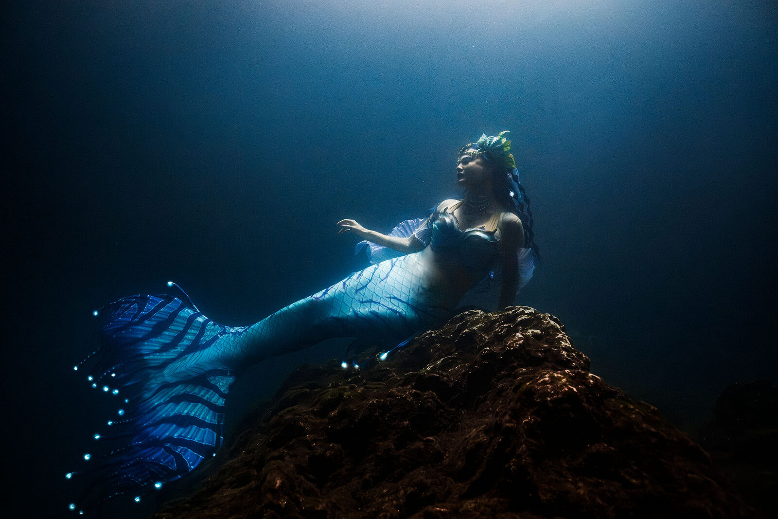 Mermaid with an iridescent blue tail reclining on a rock under a beam of light inside Devil's Den