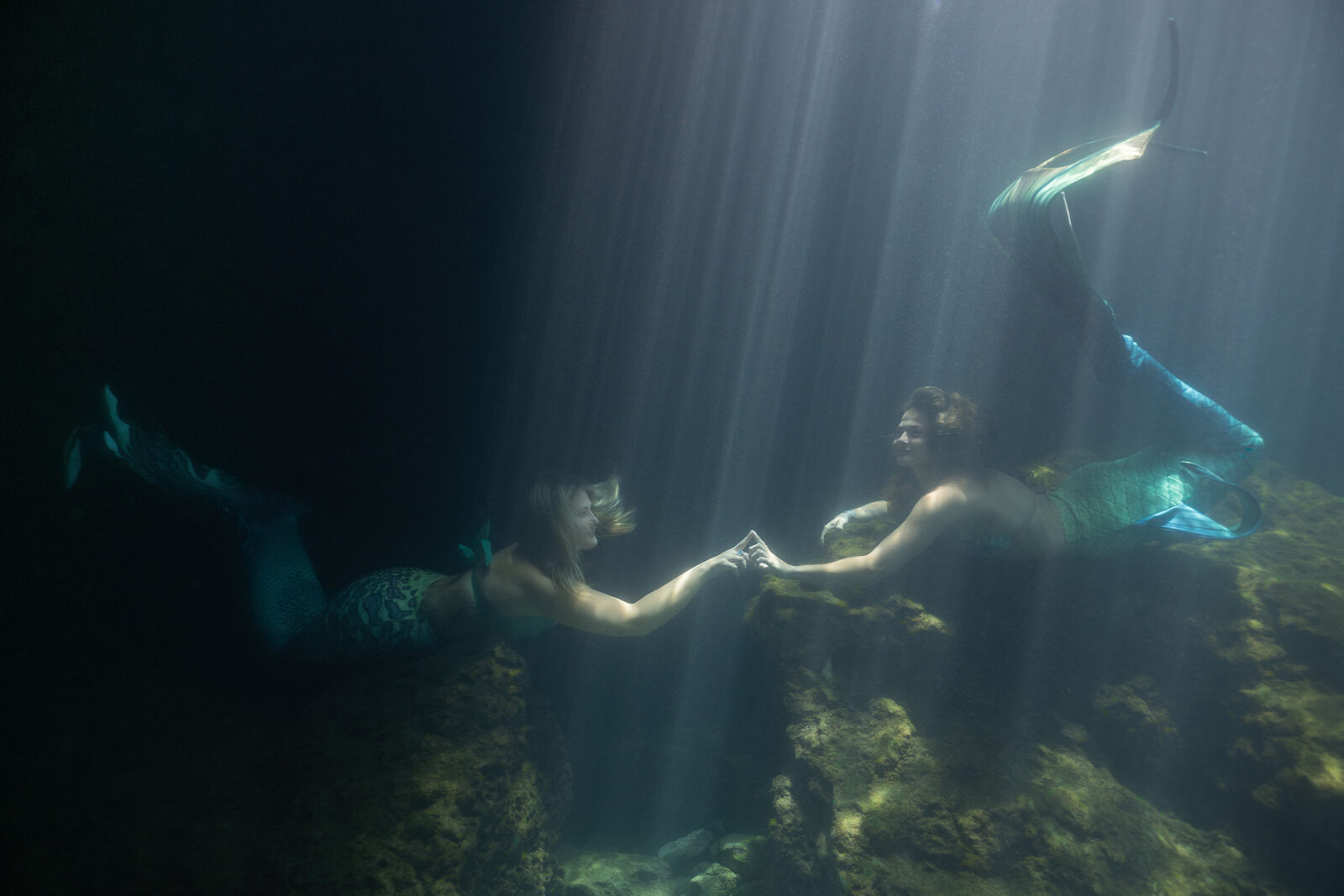 Two mermaids holding hands beneath light rays during a session at Devil's Den