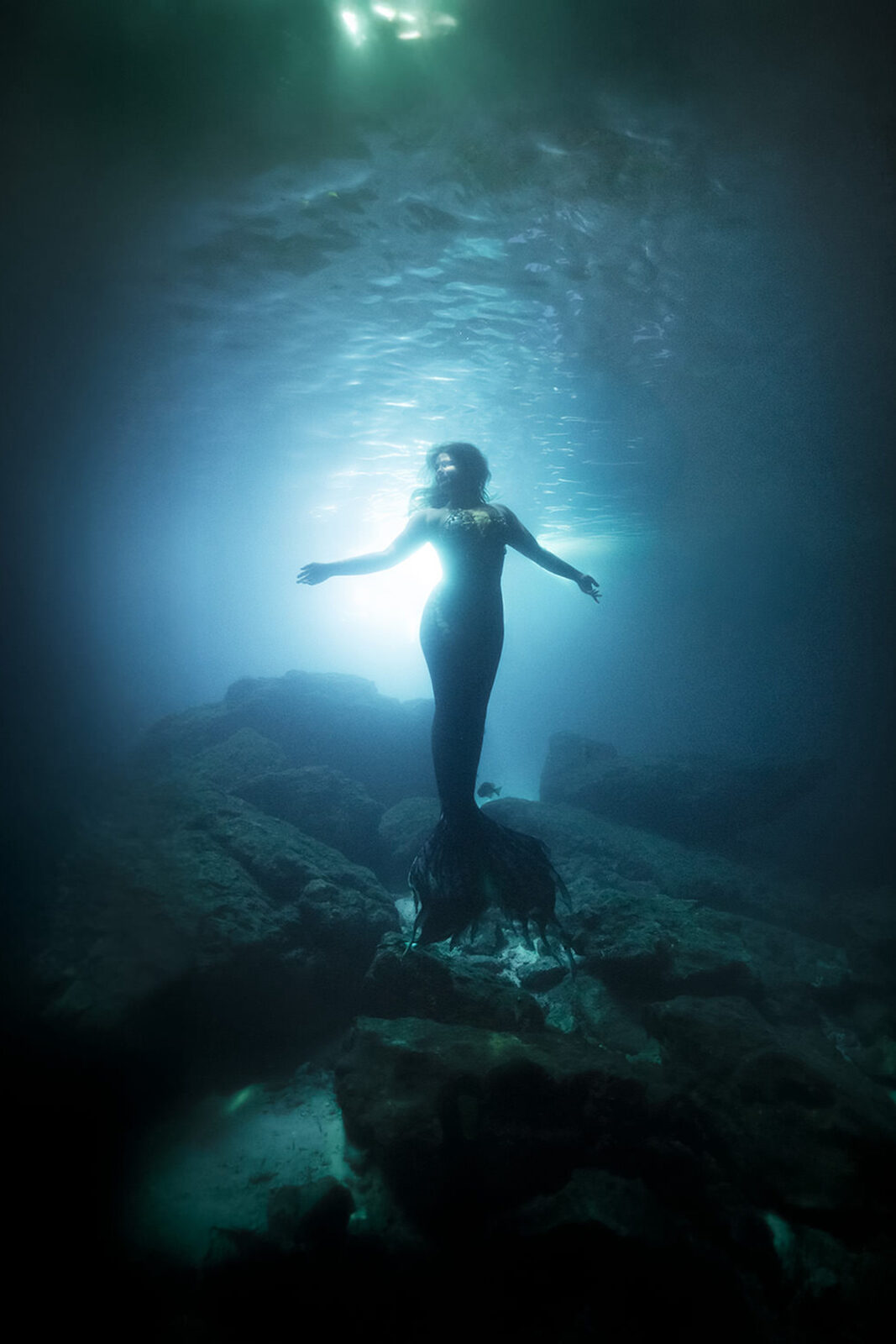Mermaid with arms spread, backlit by the blue glow of the Devil's Den cavern