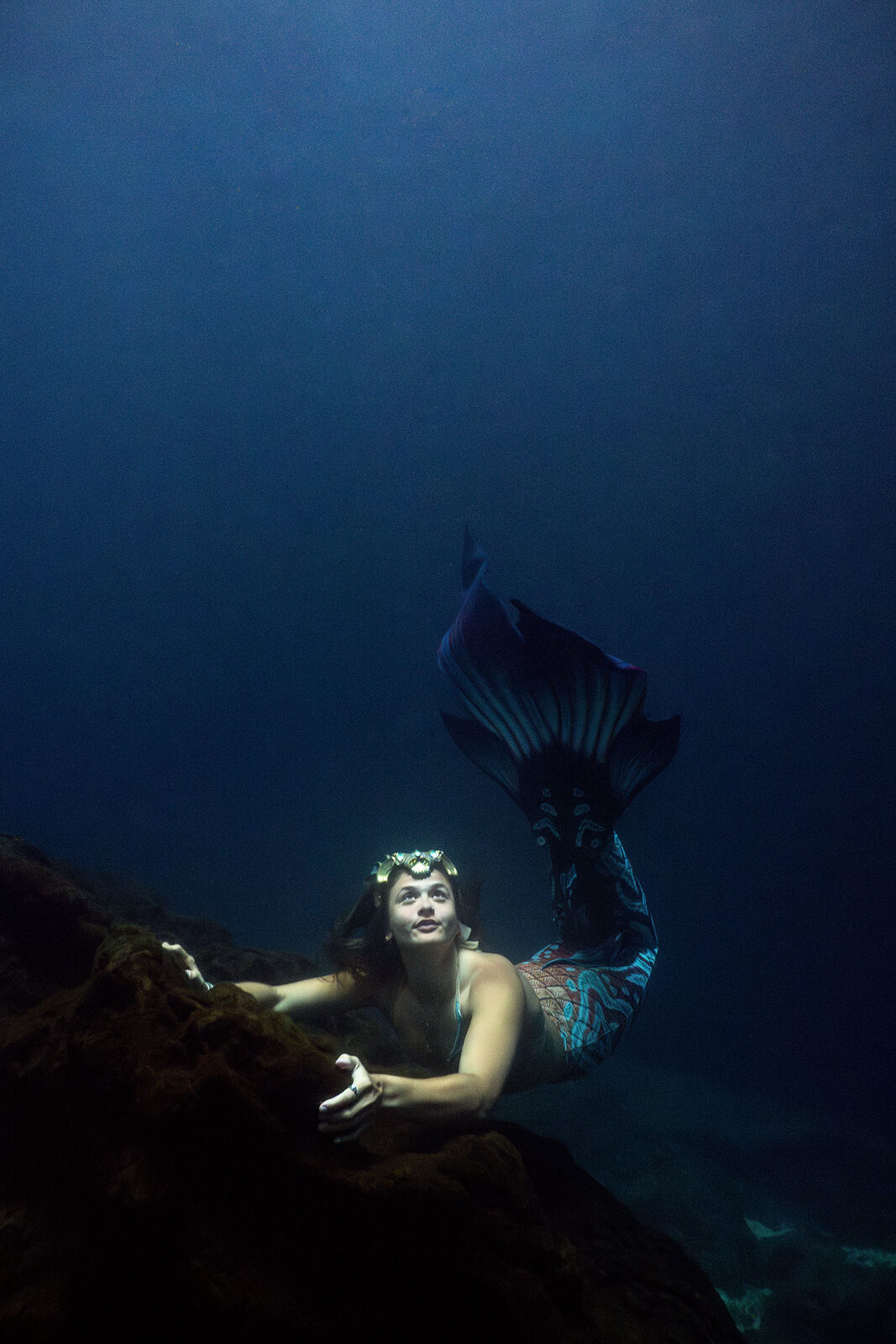 Mermaid in a blue tail and headpiece resting on a rock ledge in Devil's Den Spring